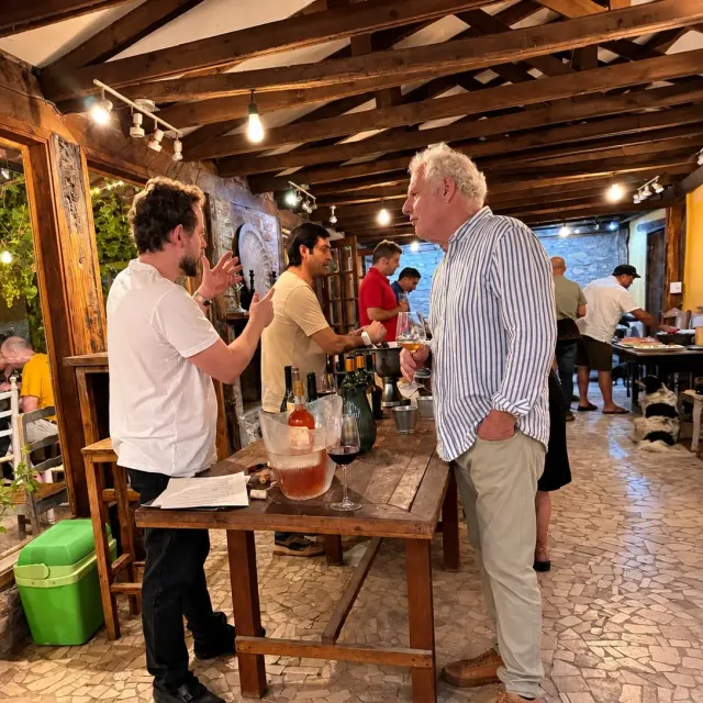 Conversación junto a una mesa de degustación con botellas y copas, bajo vigas de madera y luces cálidas